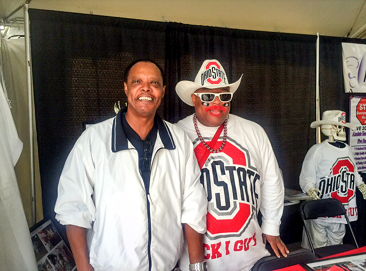 Two men pose together at a booth; one wears a white jacket, the other a white Ohio State-themed outfit with a hat and sunglasses.