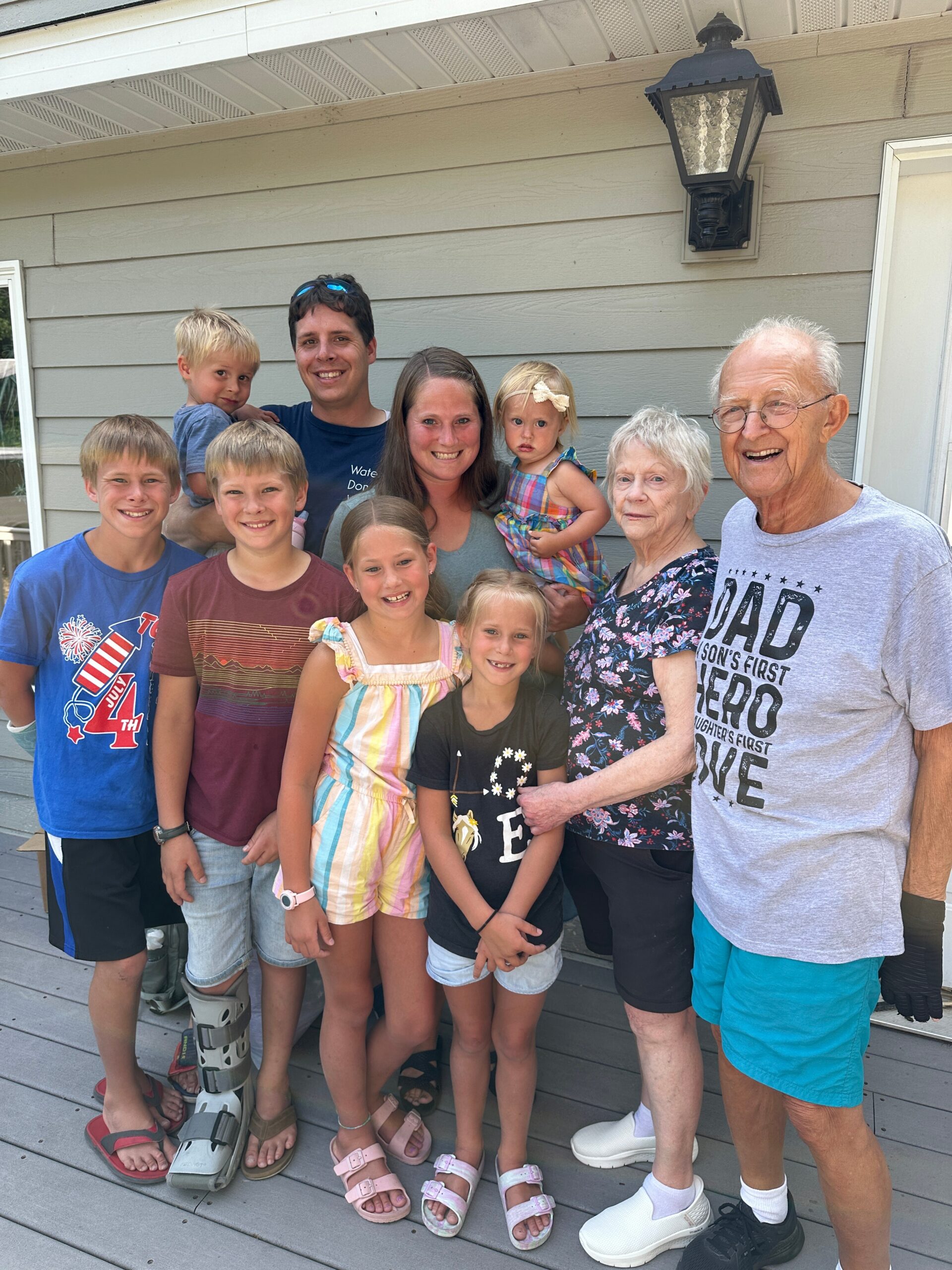 Smiling family group photo on a wooden deck in front of a grey house. Everyone faces the camera.