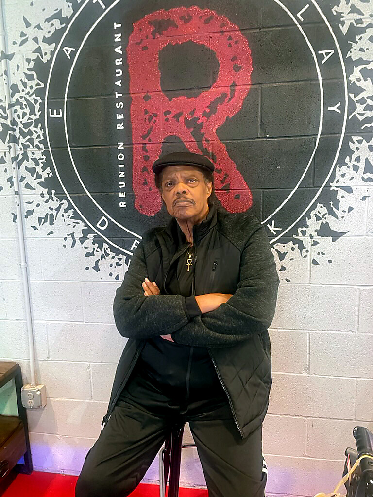 Older man in a black vest and cap sits with arms crossed in front of a wall mural featuring a large red R within a circle.