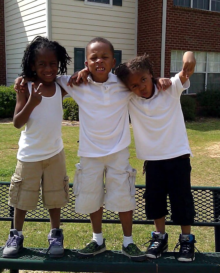Three kids with arms around each other posing on a park bench outdoors in white shirts and shorts.