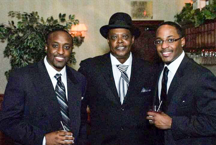 Three men in black suits pose together at a formal event, one wearing a hat, each holding a drink in a lounge setting.