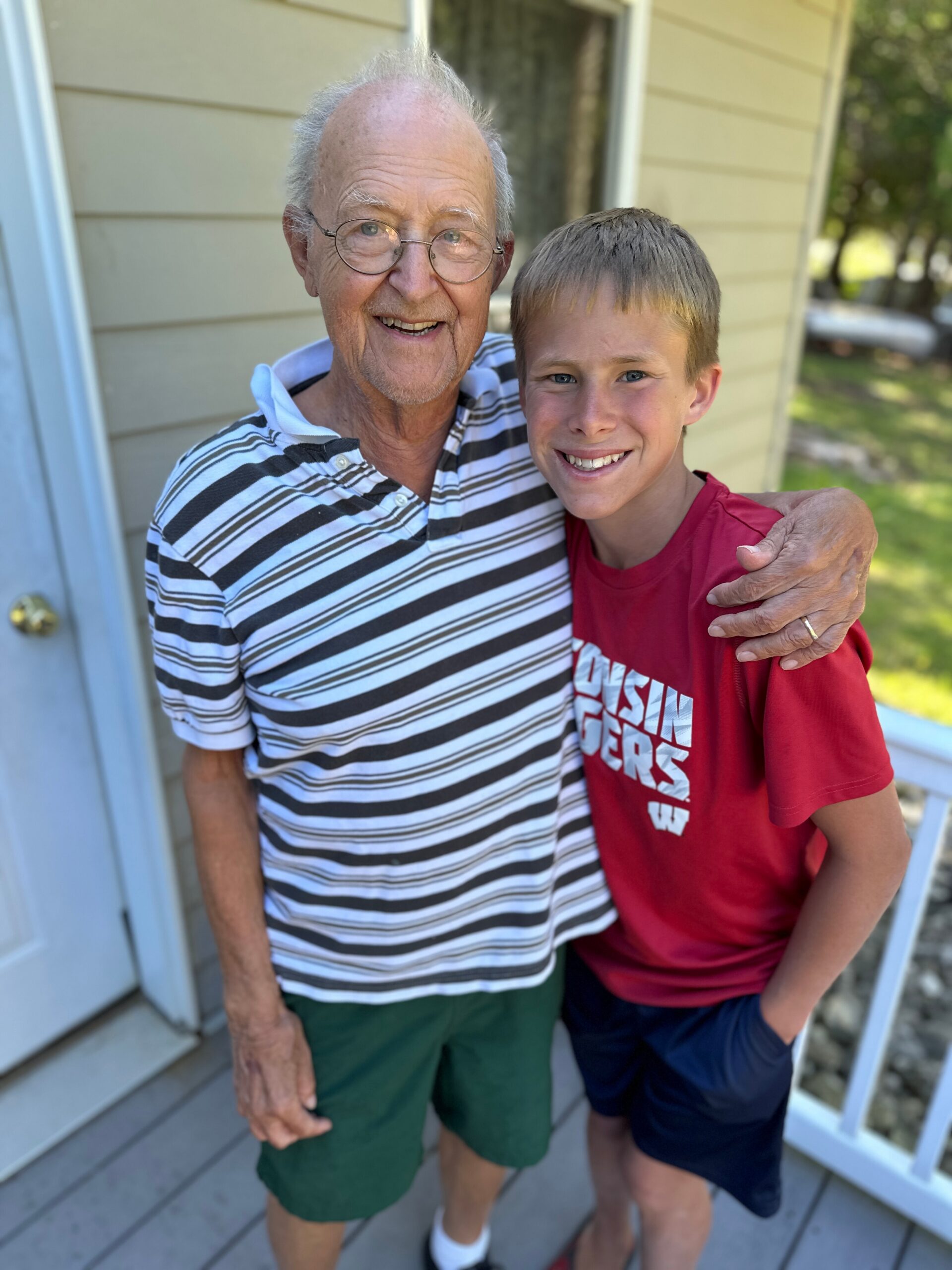 Grandfather and grandson smiling at the camera on a porch, wearing a striped shirt and red T-shirt.
