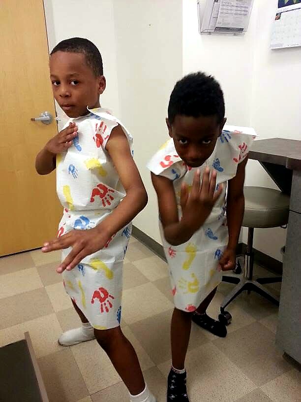 Two young children in hospital gowns pose as if dancing in a clinic, one hand extended toward the camera and the other touching their chest.