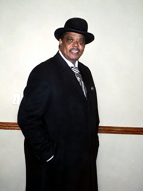 Man in a black suit and fedora posing indoors, hands in pockets and smiling.|informative image of a man in formal attire