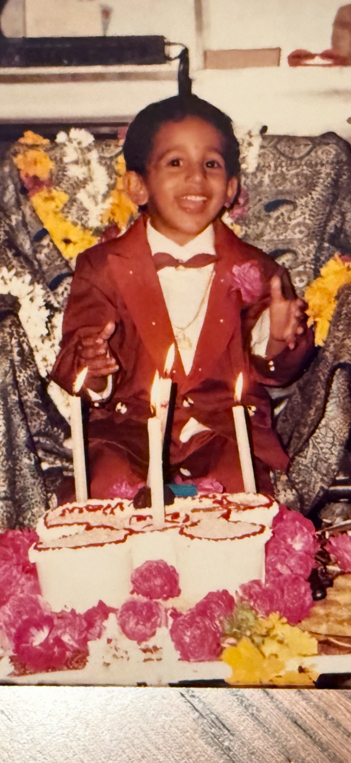 Young child in a brown suit with a bow tie sits behind a white birthday cake with three lit candles, surrounded by flowers.