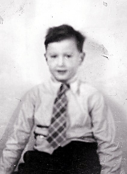 Black-and-white photo of a young boy in a dress shirt and striped tie, seated and smiling at the camera.