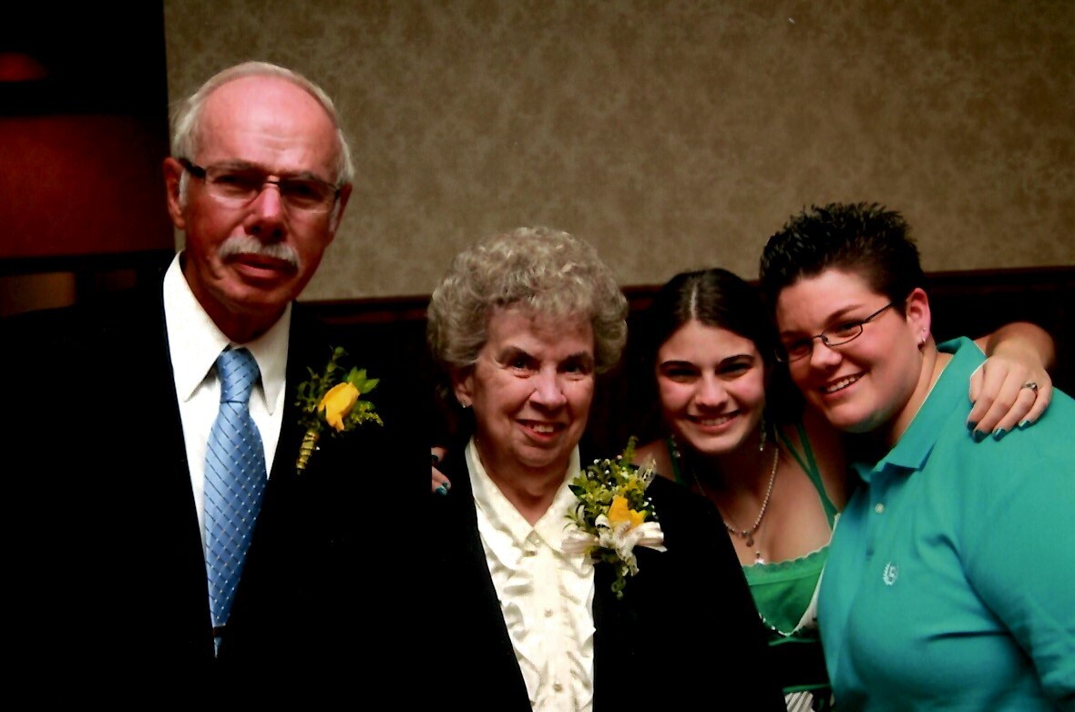 Group portrait of four smiling adults at a formal event: man in a suit with a yellow boutonniere, older woman with a corsage, younger woman in green, and a person in a turquoise polo.