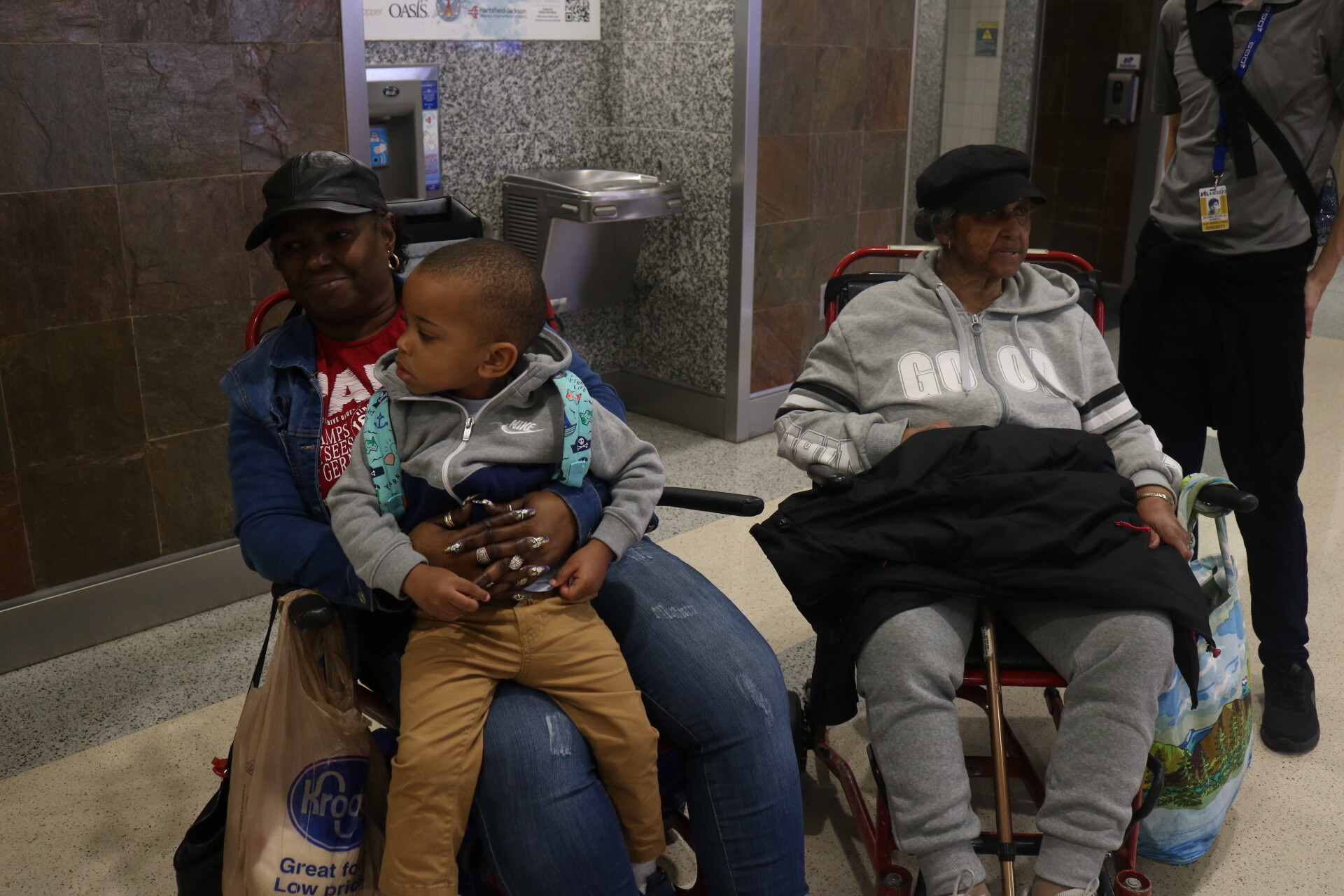 Black woman in a denim jacket sits in a wheelchair, holding a young boy in a gray hoodie on her lap; a Kroger bag hangs from her chair.