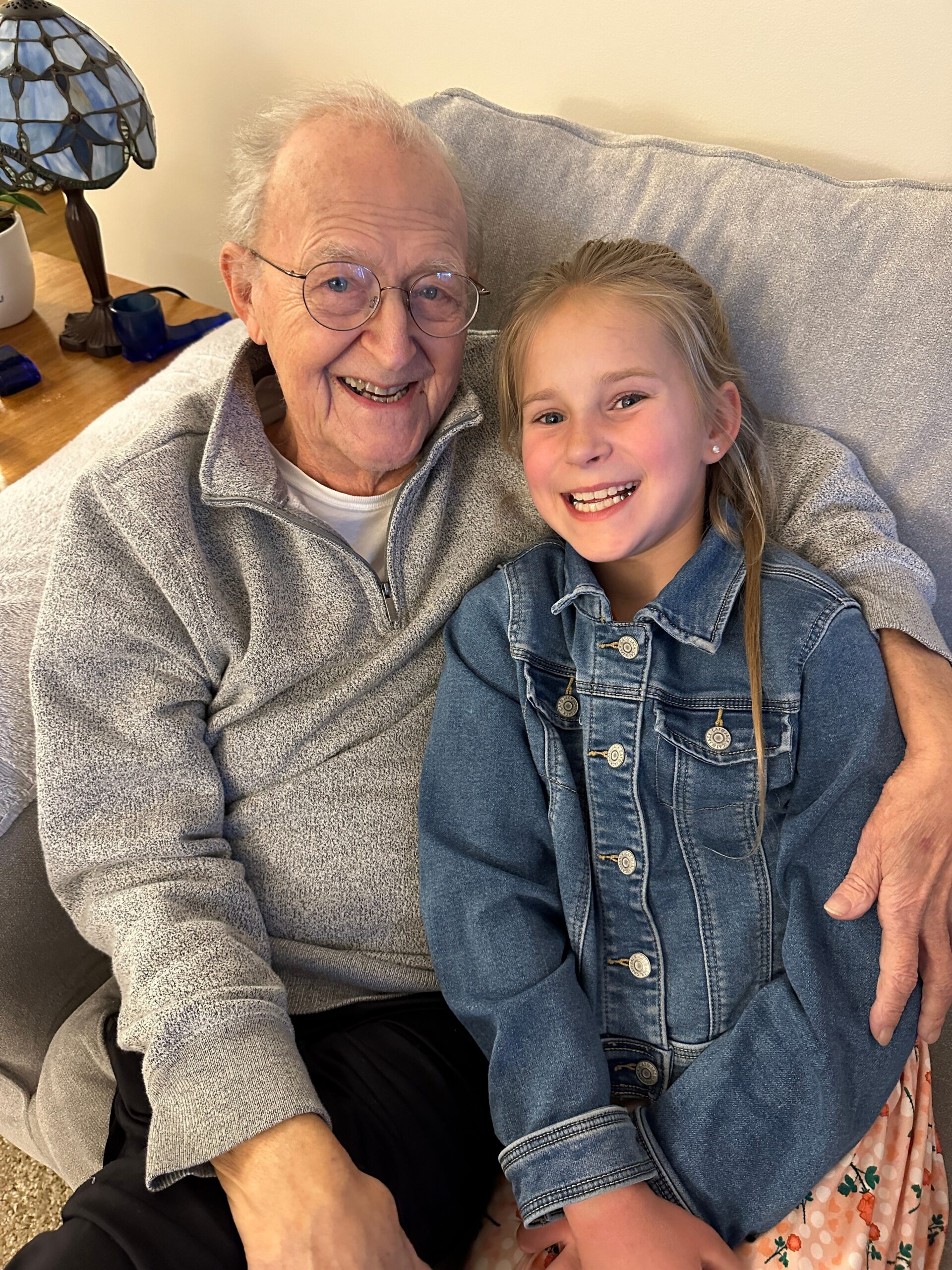 Smiling elderly man with glasses and a young girl sitting close together on a gray couch.