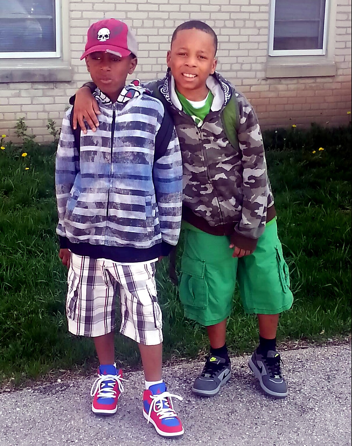 Two boys stand side by side outside a brick house, one with his arm around the other's shoulder.