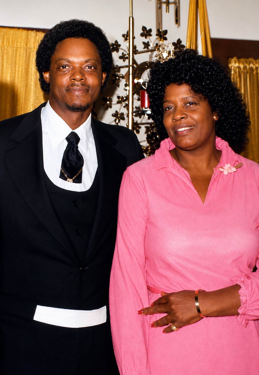 Smiling man in a black tuxedo and white bib collar standing next to a smiling woman in a pink dress at a formal event, with gold decor in the background.