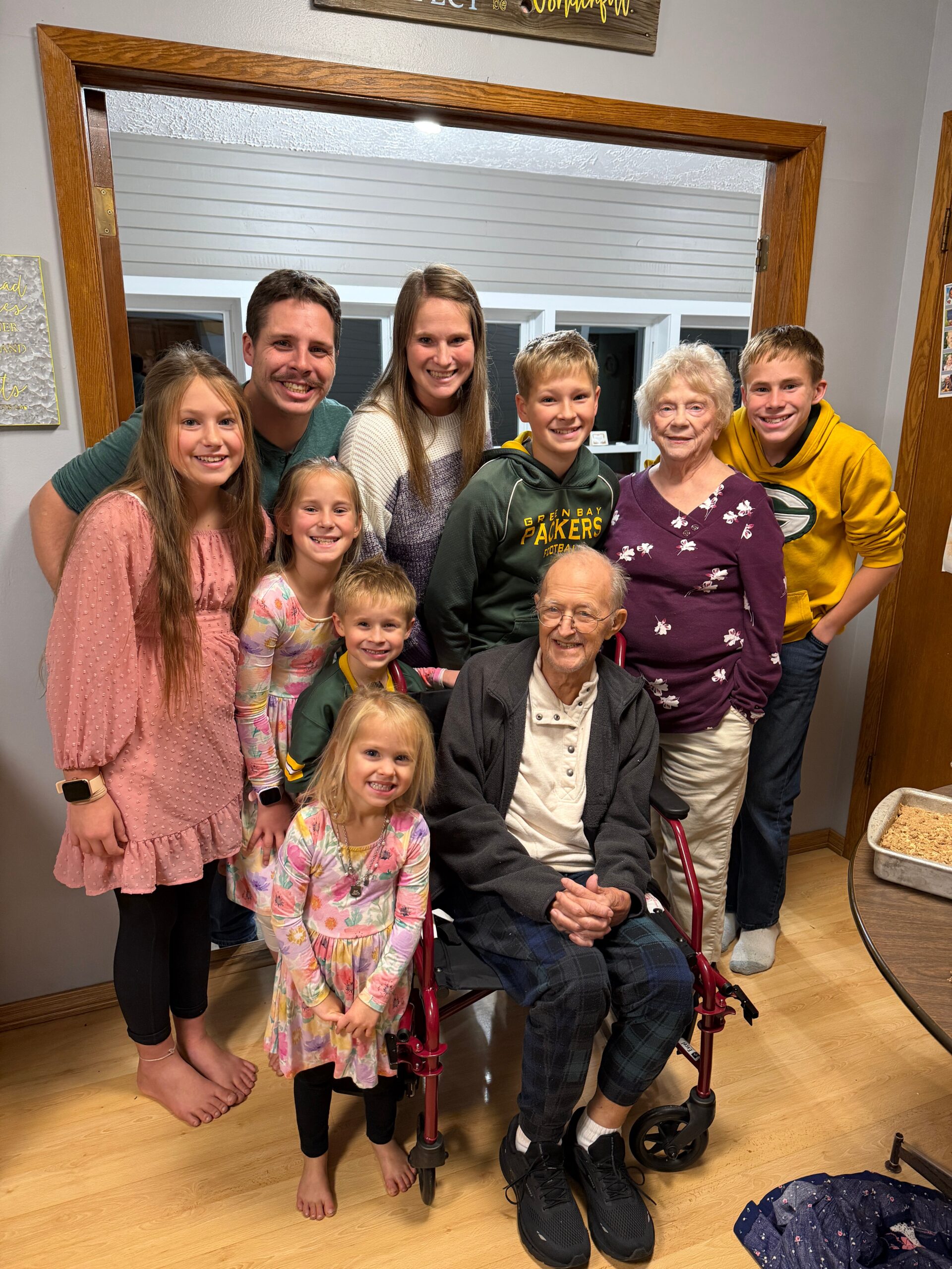 Smiling family group around an elderly man in a wheelchair indoors near a doorway.