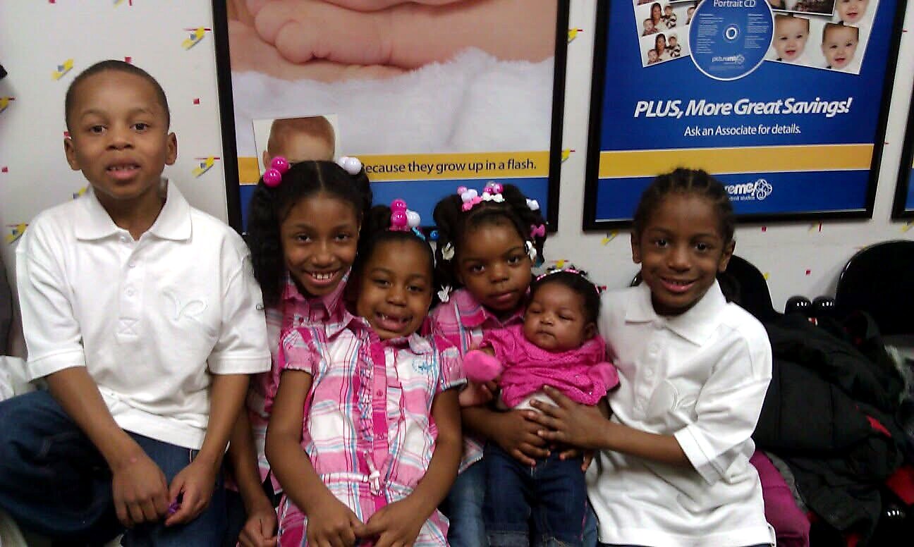 Group of children posing together in a waiting area, with colorful posters on the wall behind them and one baby in pink being held by a girl.