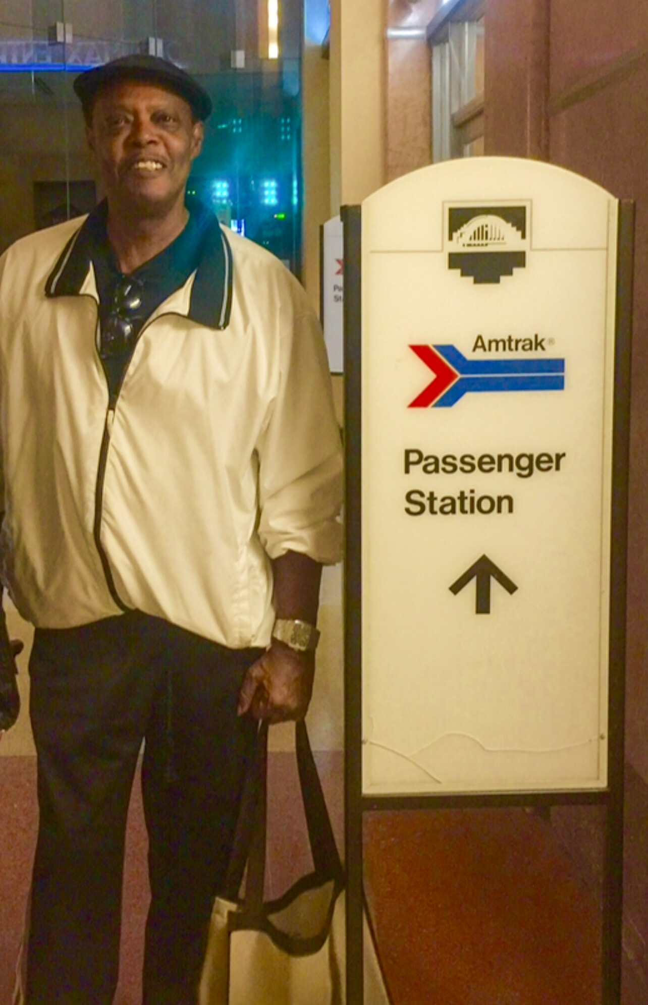 Smiling man in a beige jacket and cap standing beside an Amtrak Passenger Station sign with an upward arrow.