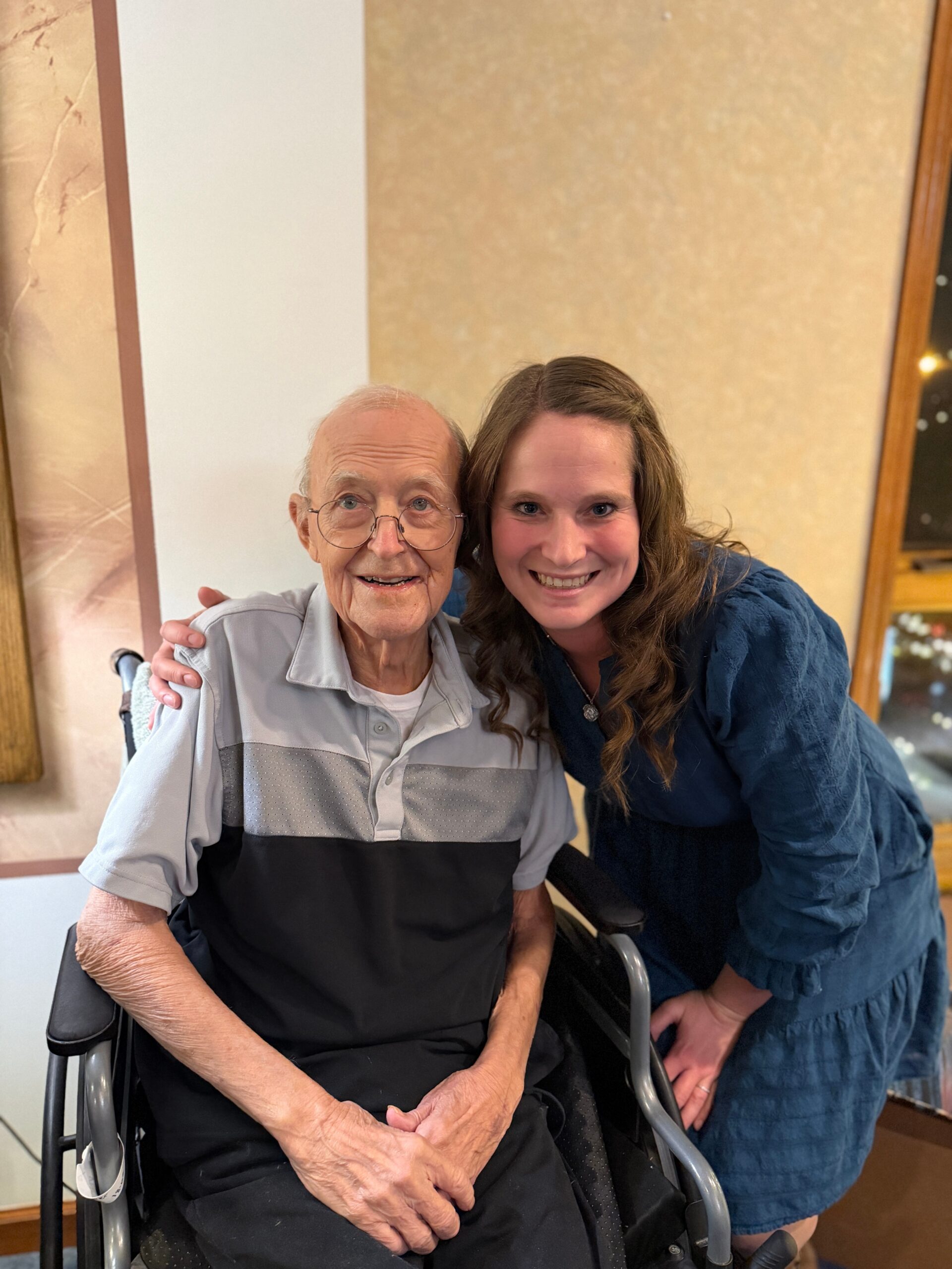 Smiling elderly man in a wheelchair with a younger woman leaning in for a hug beside him.