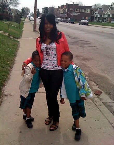 Woman with two young boys standing on a sidewalk in a suburban street, arms around each other.