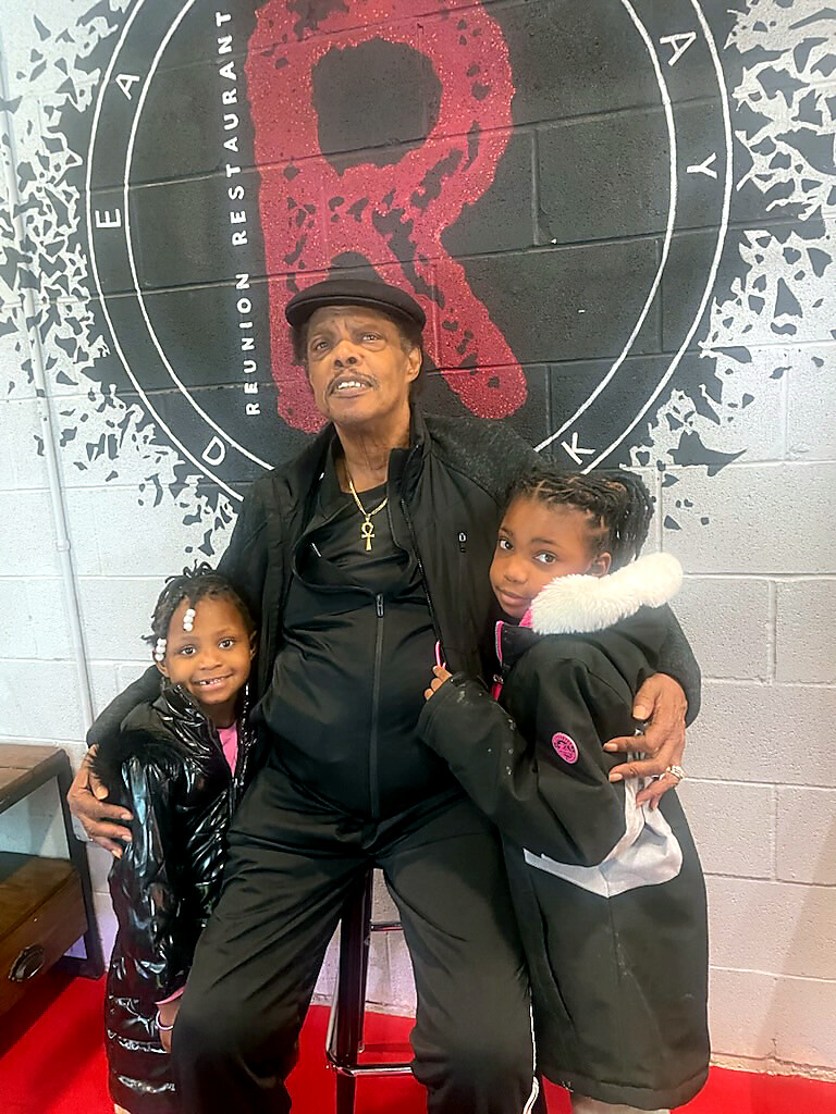 A man in a black outfit and hat sits on a stool between two smiling young girls, posing together in front of a wall with a large circular logo behind them.