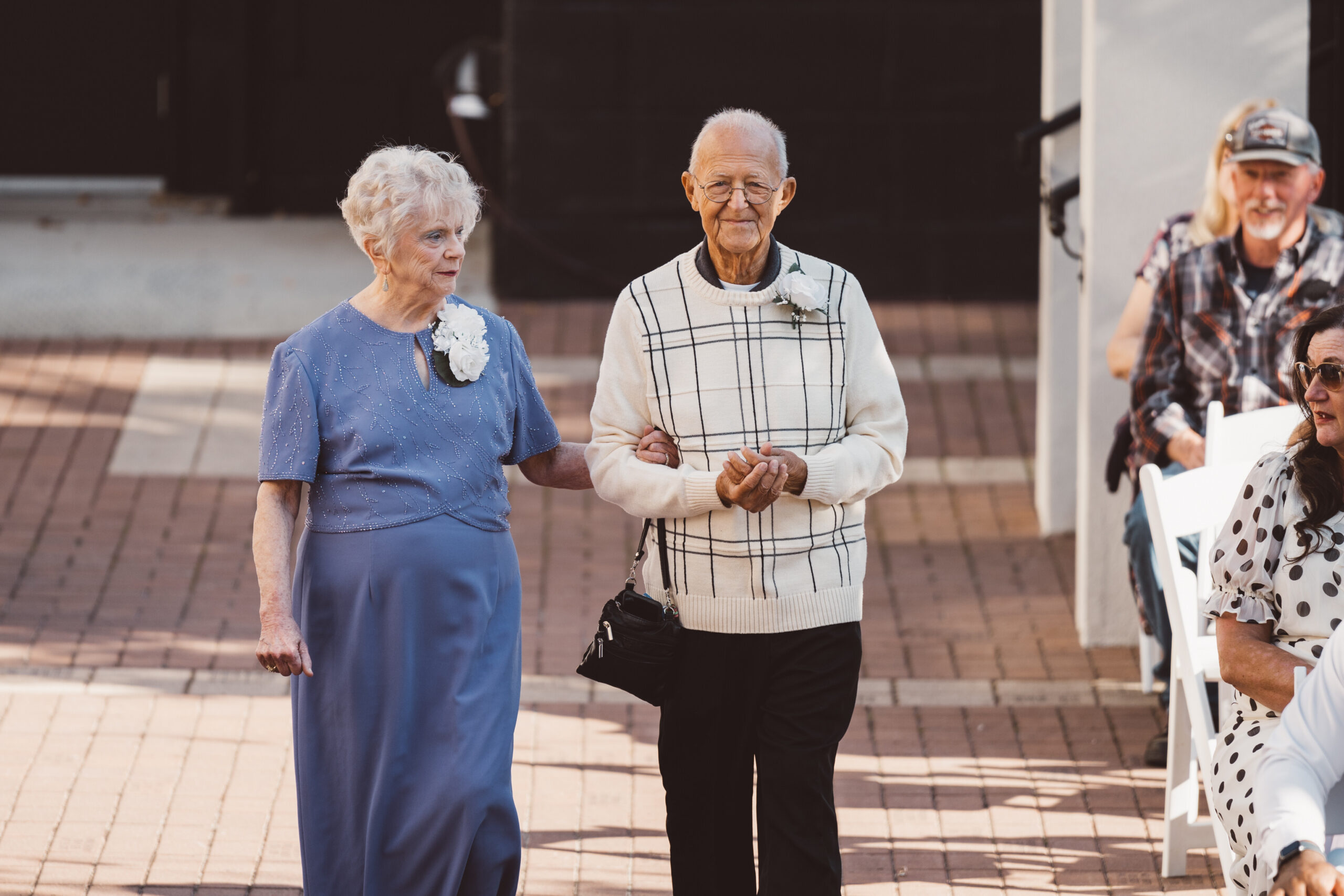 Elderly woman in a blue dress and elderly man in a white plaid sweater walk arm in arm at an outdoor event, with spectators on the right.