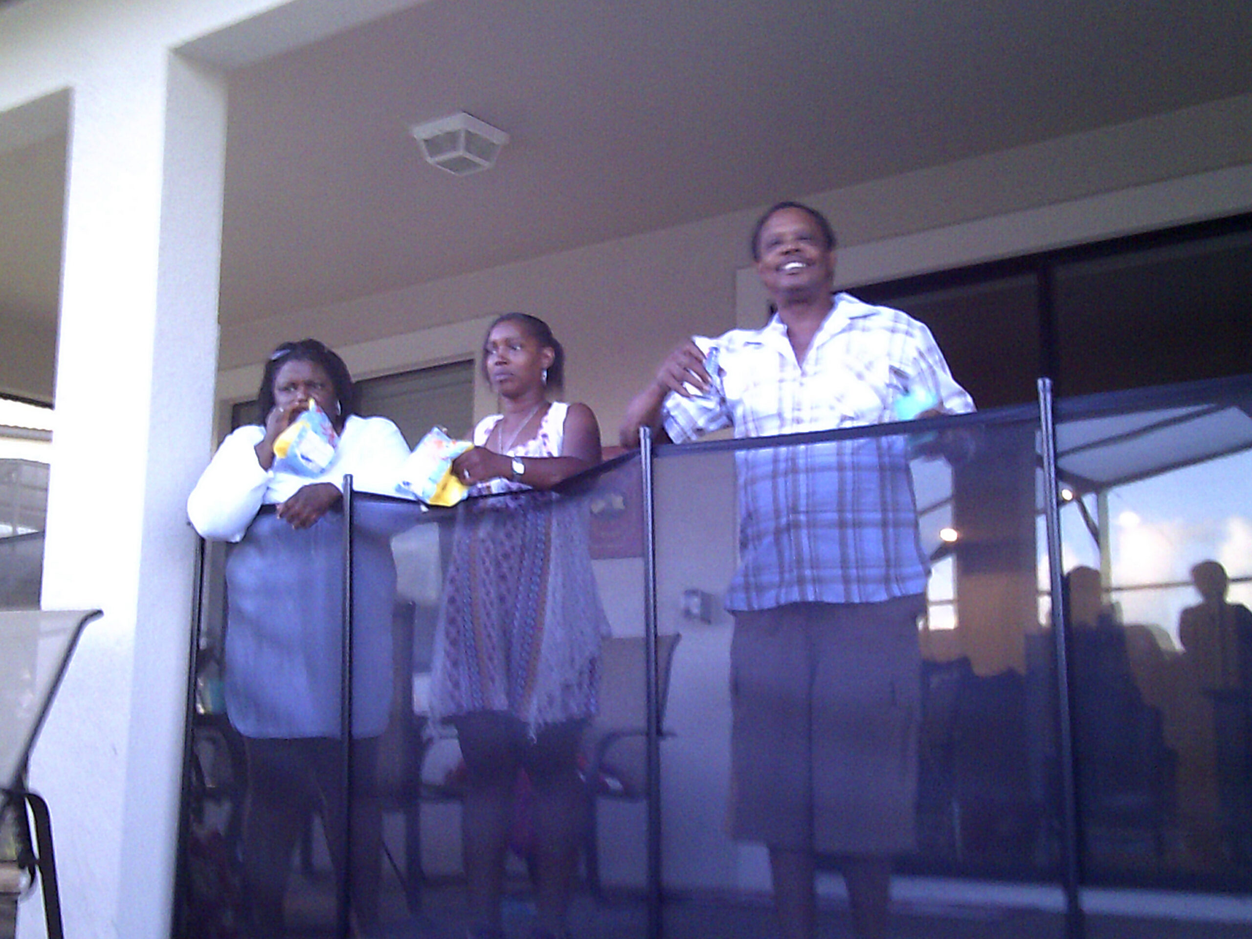 Group of three people standing on a balcony, leaning on a glass railing with snacks in hand, smiling and chatting.