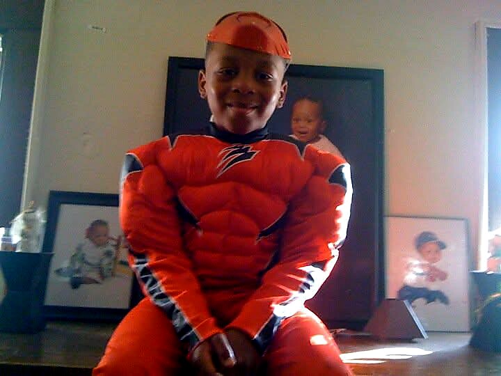 Smiling child in an orange superhero costume kneeling indoors on the floor of a home.