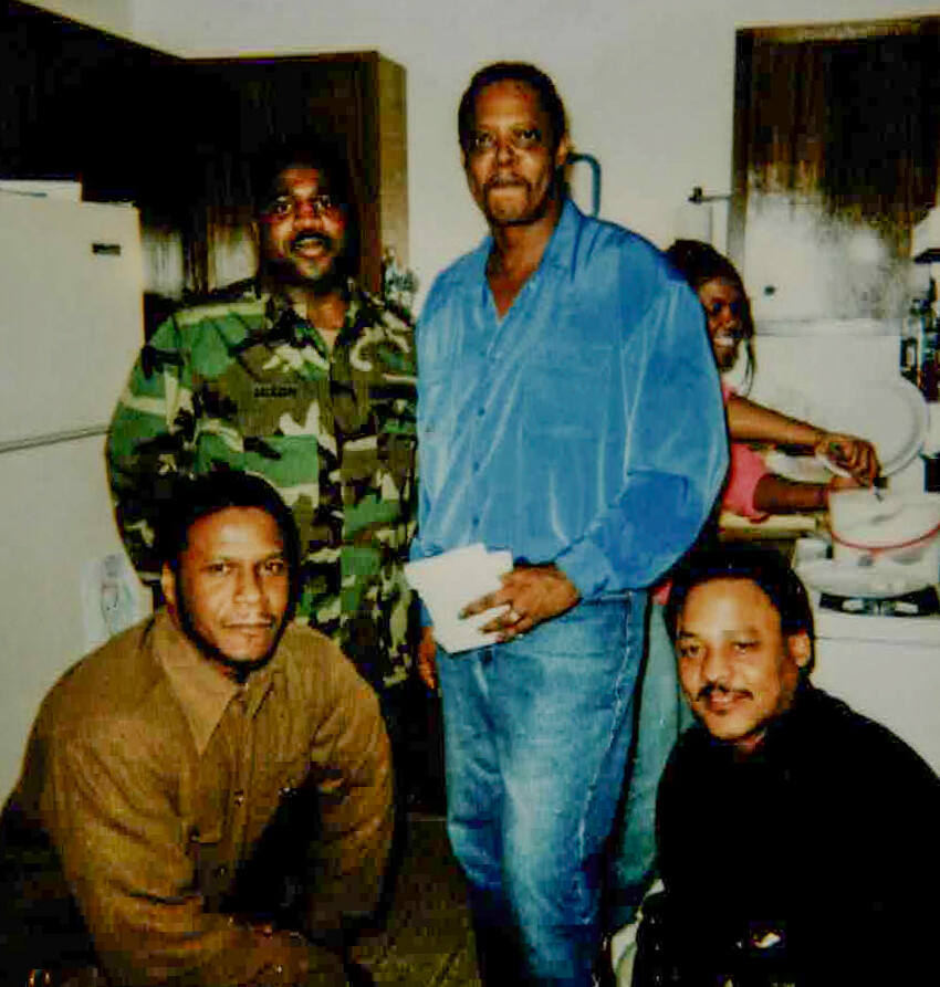 Five people in a home laundry room: two men seated on the floor, one in camouflage jacket, another in brown shirt, and a man in blue pajama-style clothing standing and holding papers, with a woman at a washing machine in the background.