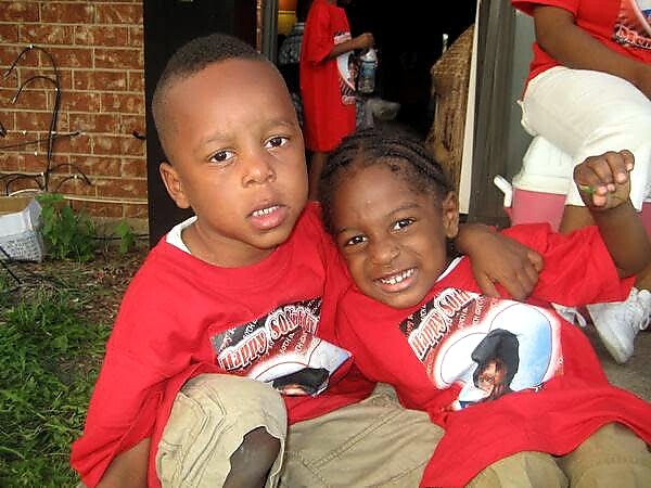 Two young children sit closely together outdoors, wearing matching red shirts, smiling with their arms around each other against a brick wall backdrop.