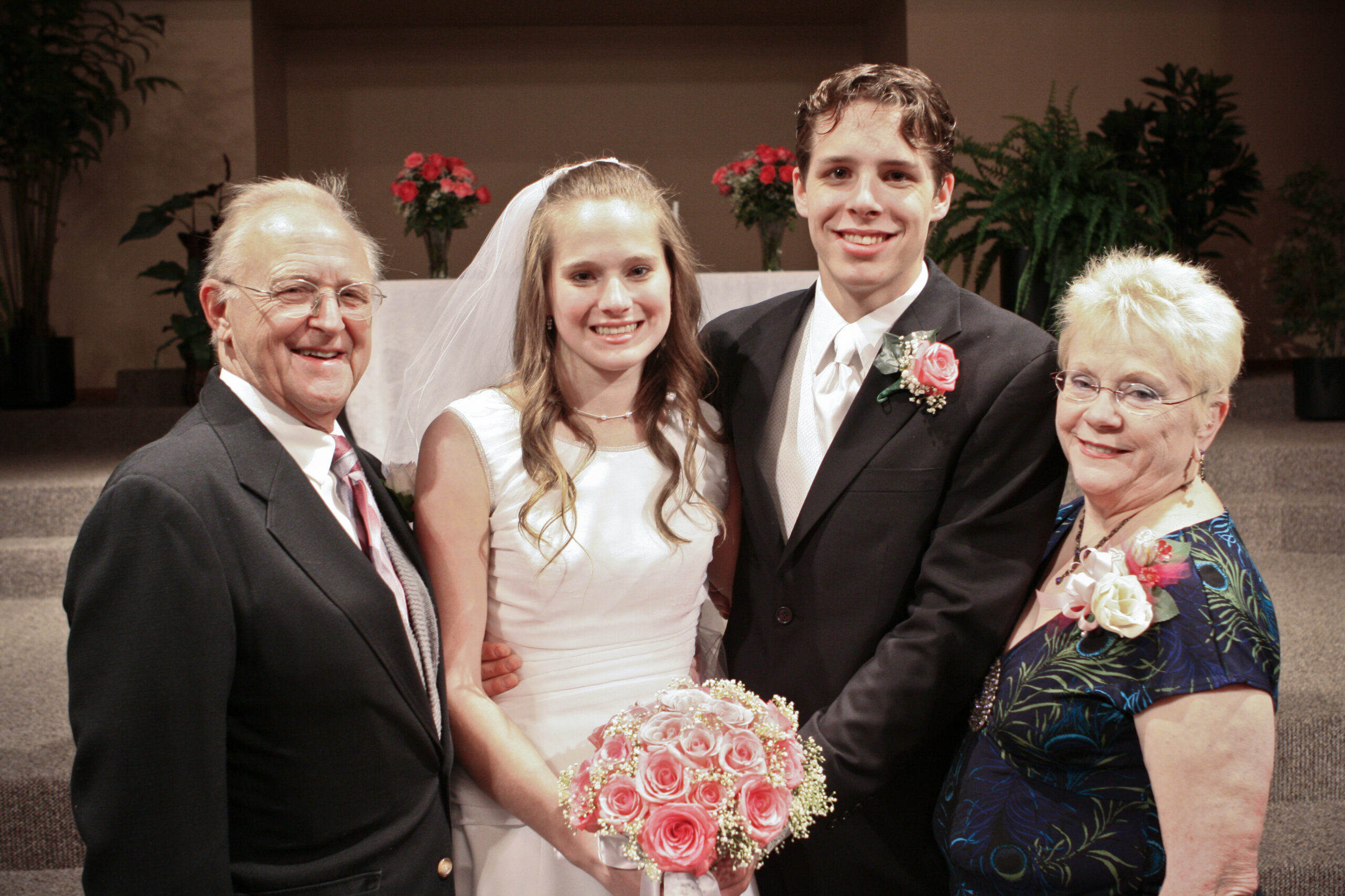 Bride and groom posing with smiling parents, indoors wedding setting; bride holds pink rose bouquet.