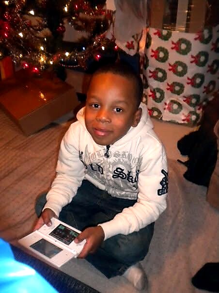 A young boy sits on a living room rug by a Christmas tree, holding a handheld game console.