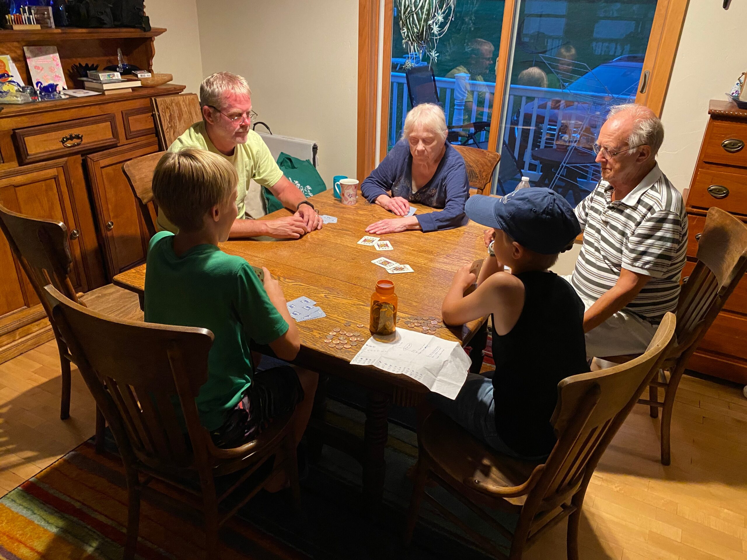 Six people— adults and children—sitting around a wooden dining table playing cards with coins and papers nearby in a cozy home dining area.