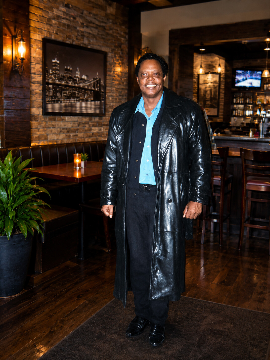 Smiling man in a shiny black trench coat and blue shirt poses in a warmly lit bar/restaurant Interior.