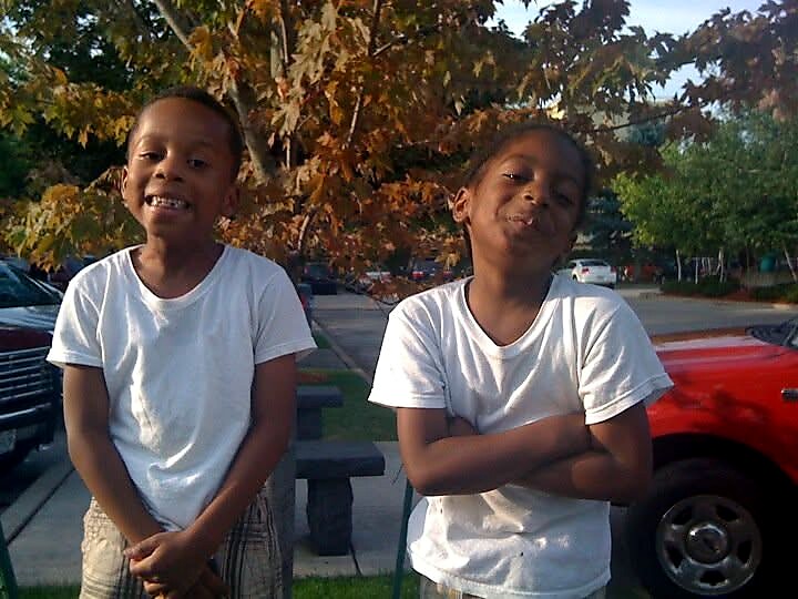 Two smiling children in white shirts pose outdoors with autumn leaves in the background.