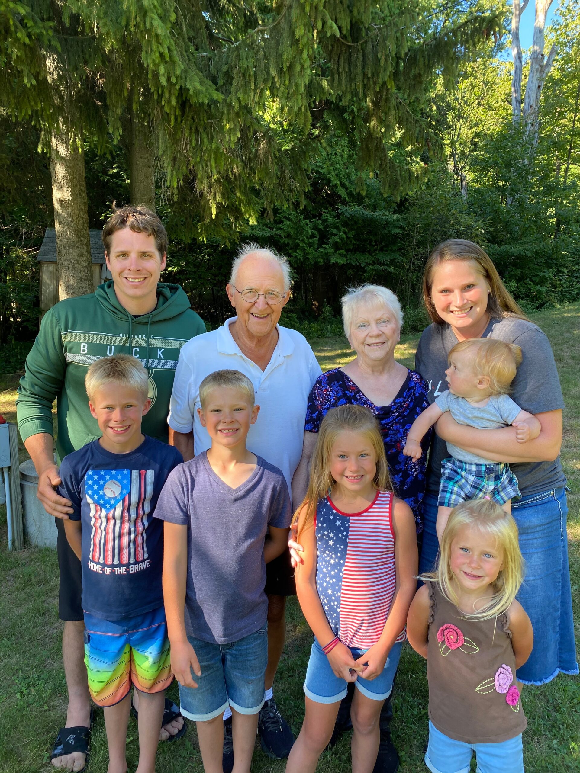 Multi-generation family posing for a photo outdoors in a sunny yard with tall trees in the background, all smiling.