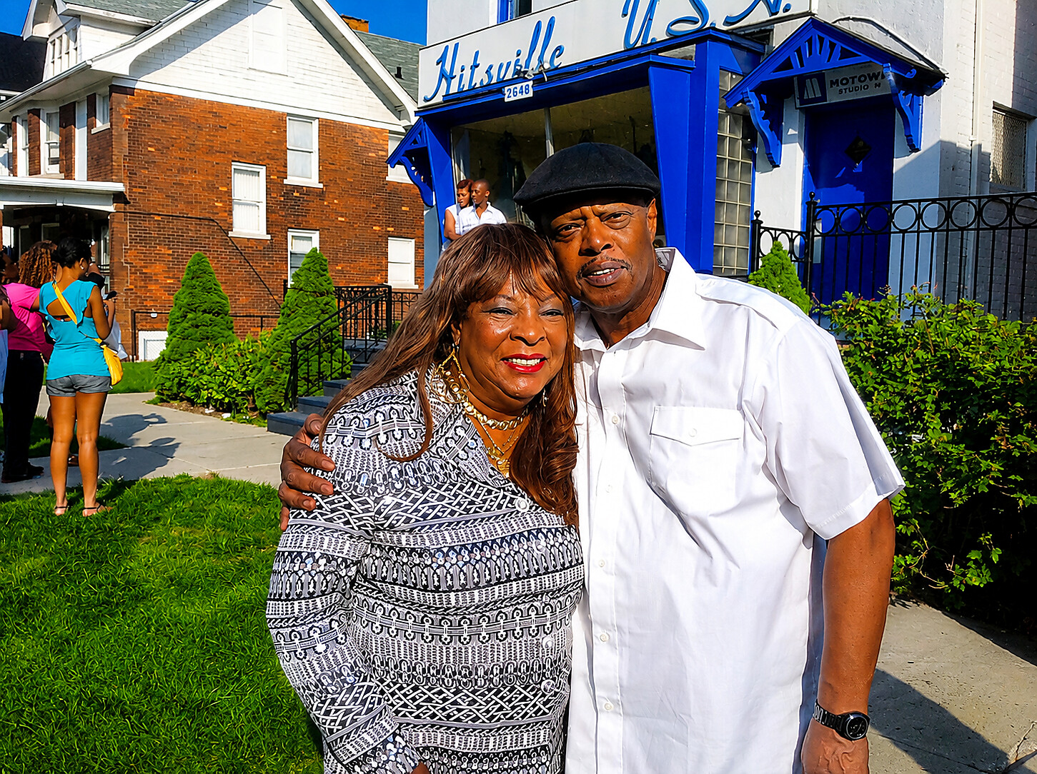 Couple poses together outside a blue-and-white building with “Hitsville U.S.A.” signage, smiling at the camera (Man in white shirt and hat, woman in patterned dress).