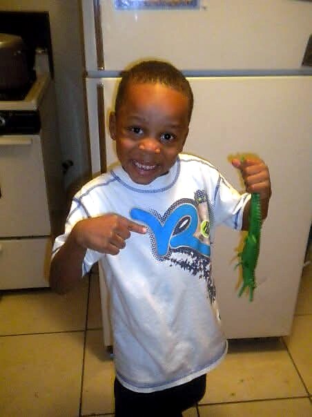 Smiling young boy in a white T-shirt points to a green plastic toy he holds in his left hand in a kitchen setting.