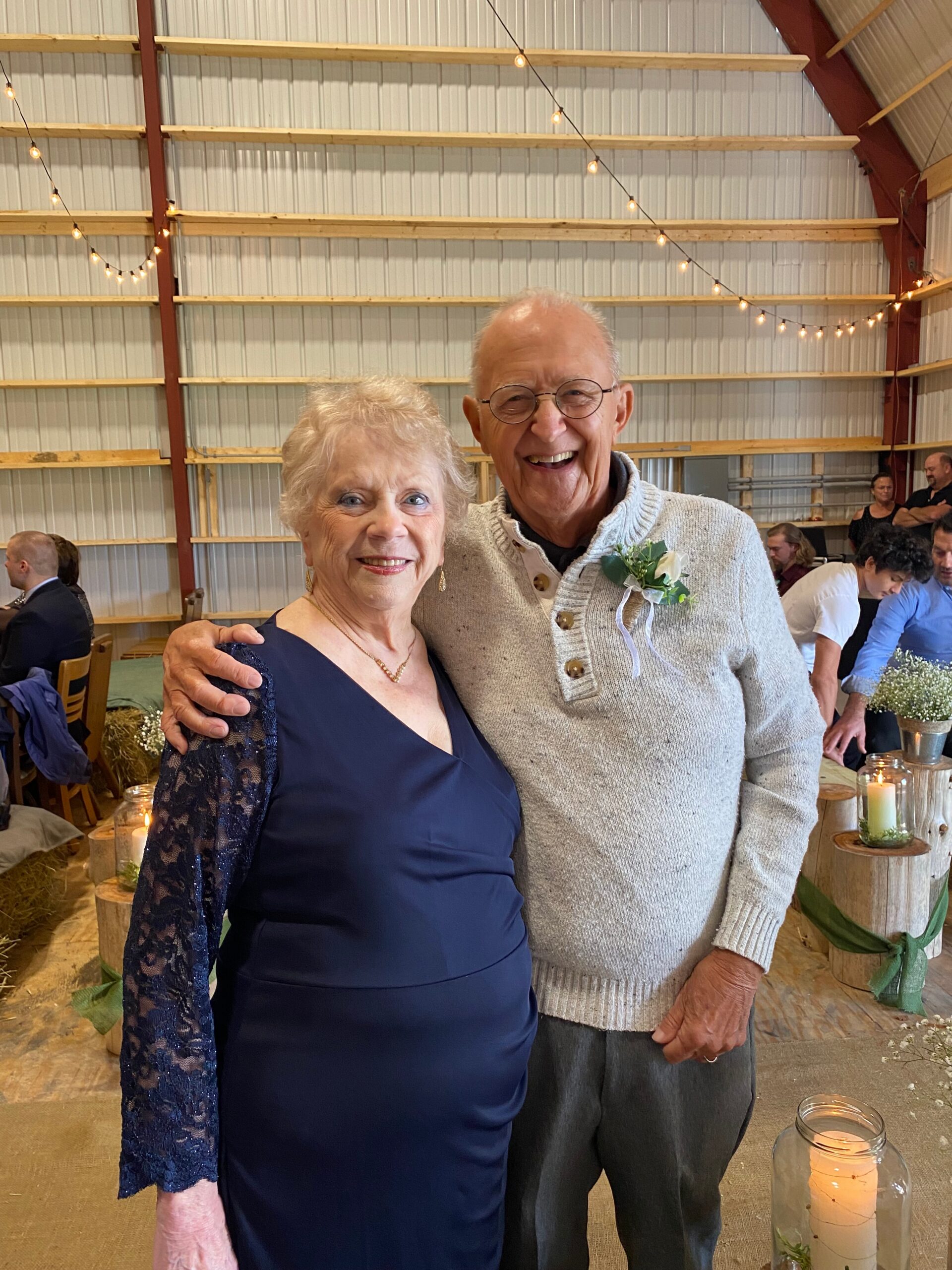 Happy elderly couple posing for a photo at a rustic indoor event, string lights overhead and candles on the tables in the background.