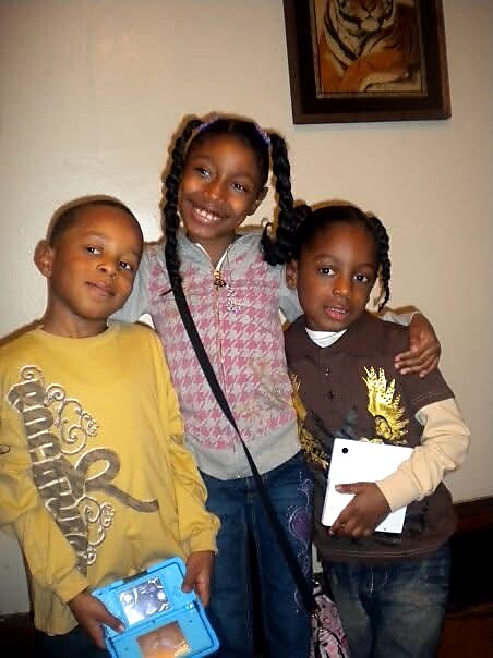 Three smiling children stand together indoors, arms around each other: left girl in yellow holds a blue device case, middle girl with braids wears pink jacket, right girl in brown holds a notebook and hugs her friends.