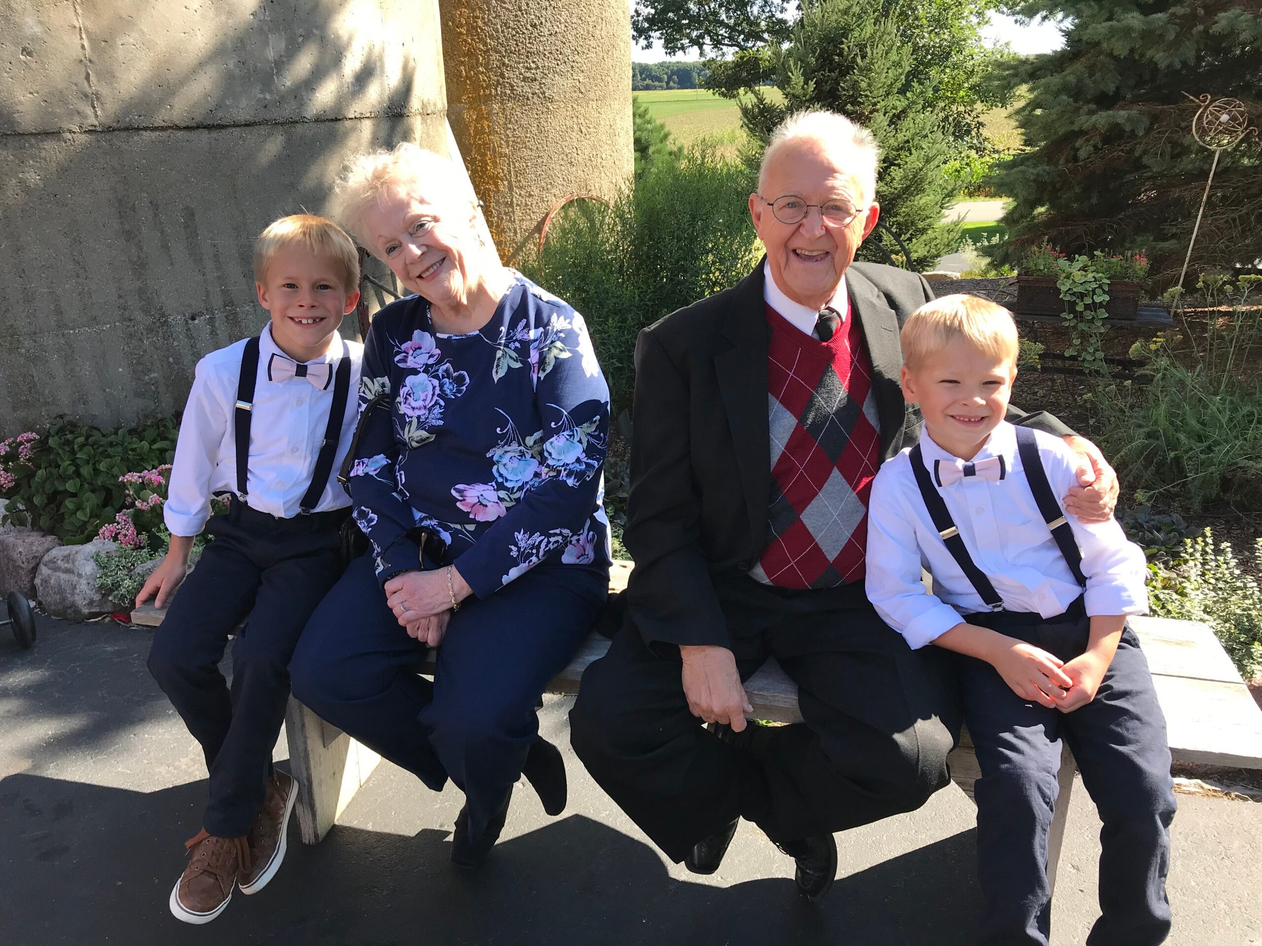 Four family members sit on a bench outdoors, two grandparents with two young boys in formal attire, all smiling at the camera.