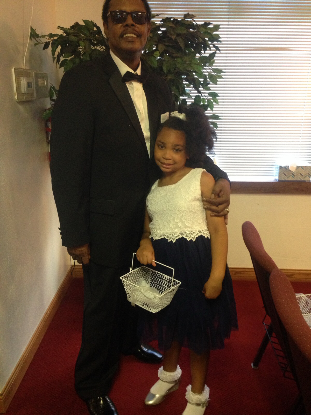 Man in black tuxedo and sunglasses posing with a young girl in a white lace dress, inside a room with blinds; girl holds a small white basket.