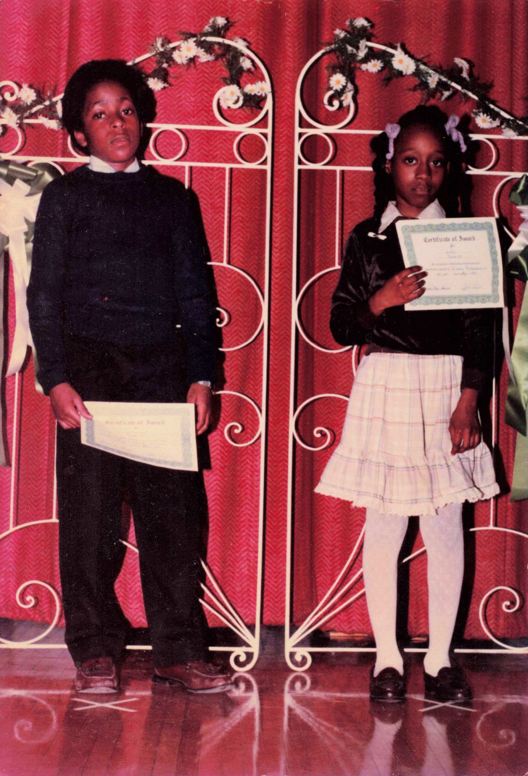 Two children stand side by side on a stage, each holding a certificate during a school awards ceremony with a red curtain backdrop and decorative white gate behind them.