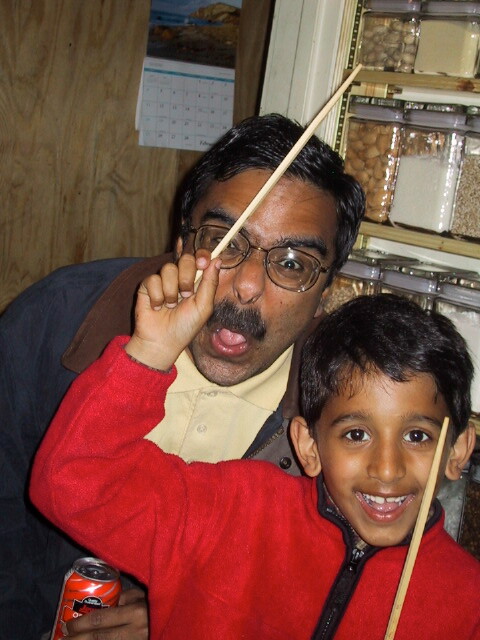 Father and young boy posing playfully with wooden sticks held near their faces in a shop.