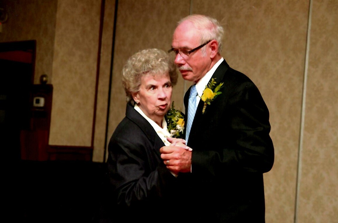 An elderly couple in formal attire dance together, wearing yellow boutonnieres at a decorated indoor event.