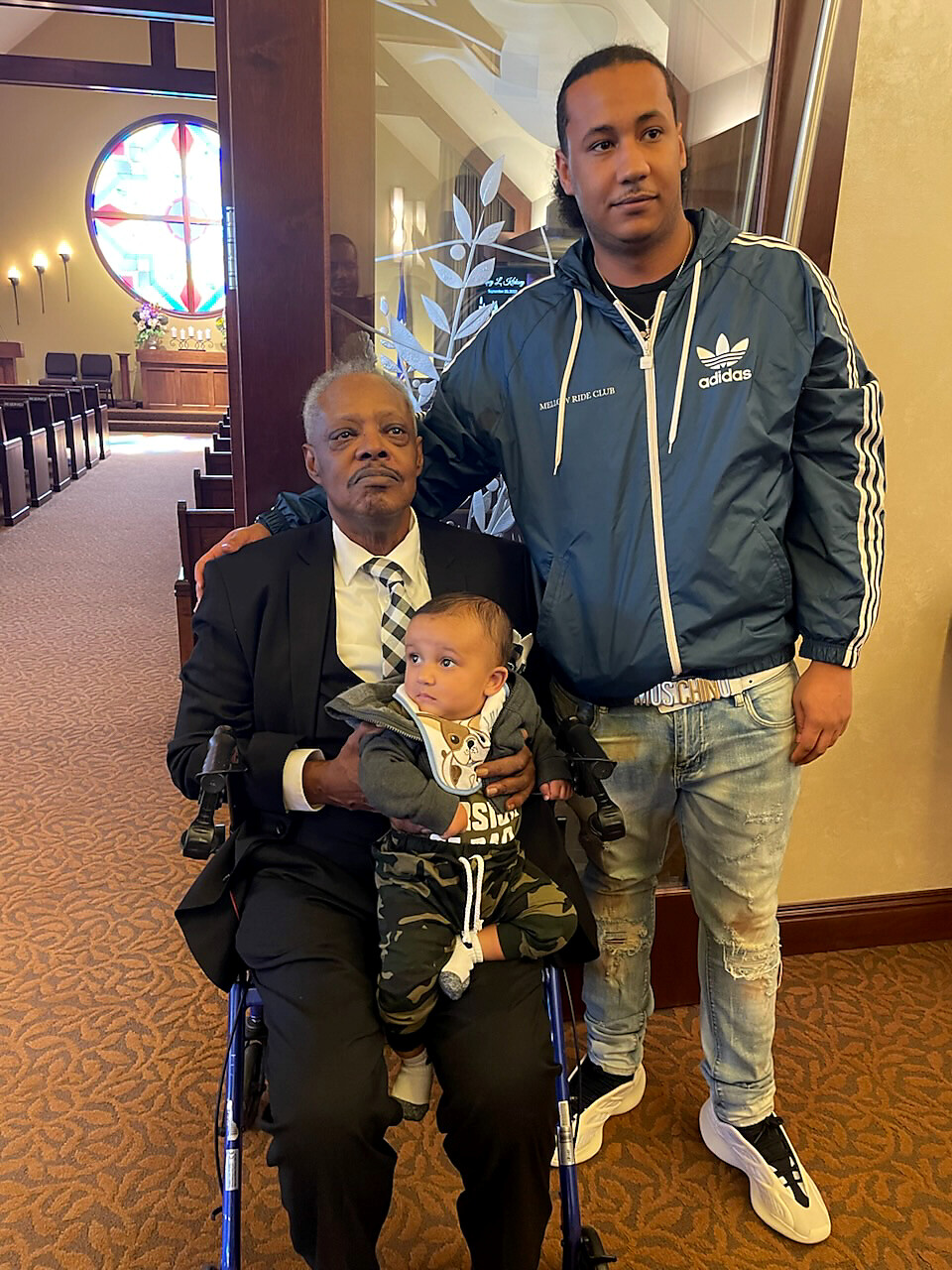 Older man in a suit seated in a wheelchair holds a baby while a younger man in a blue Adidas jacket stands beside them in a church foyer.