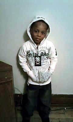 Child standing indoors against a plain wall, wearing a white hoodie and dark pants with hands in pockets.