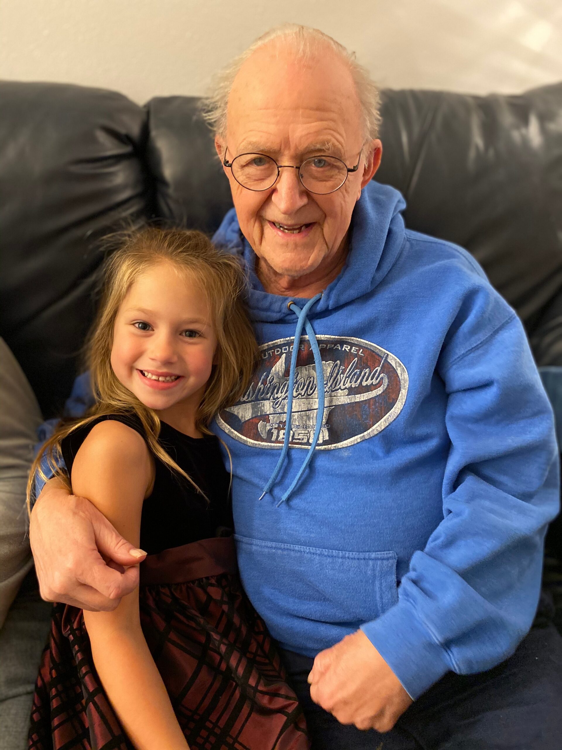 Grandfather in a blue hoodie smiling with a young girl in a black dress sitting on a black sofa together on a couch image.