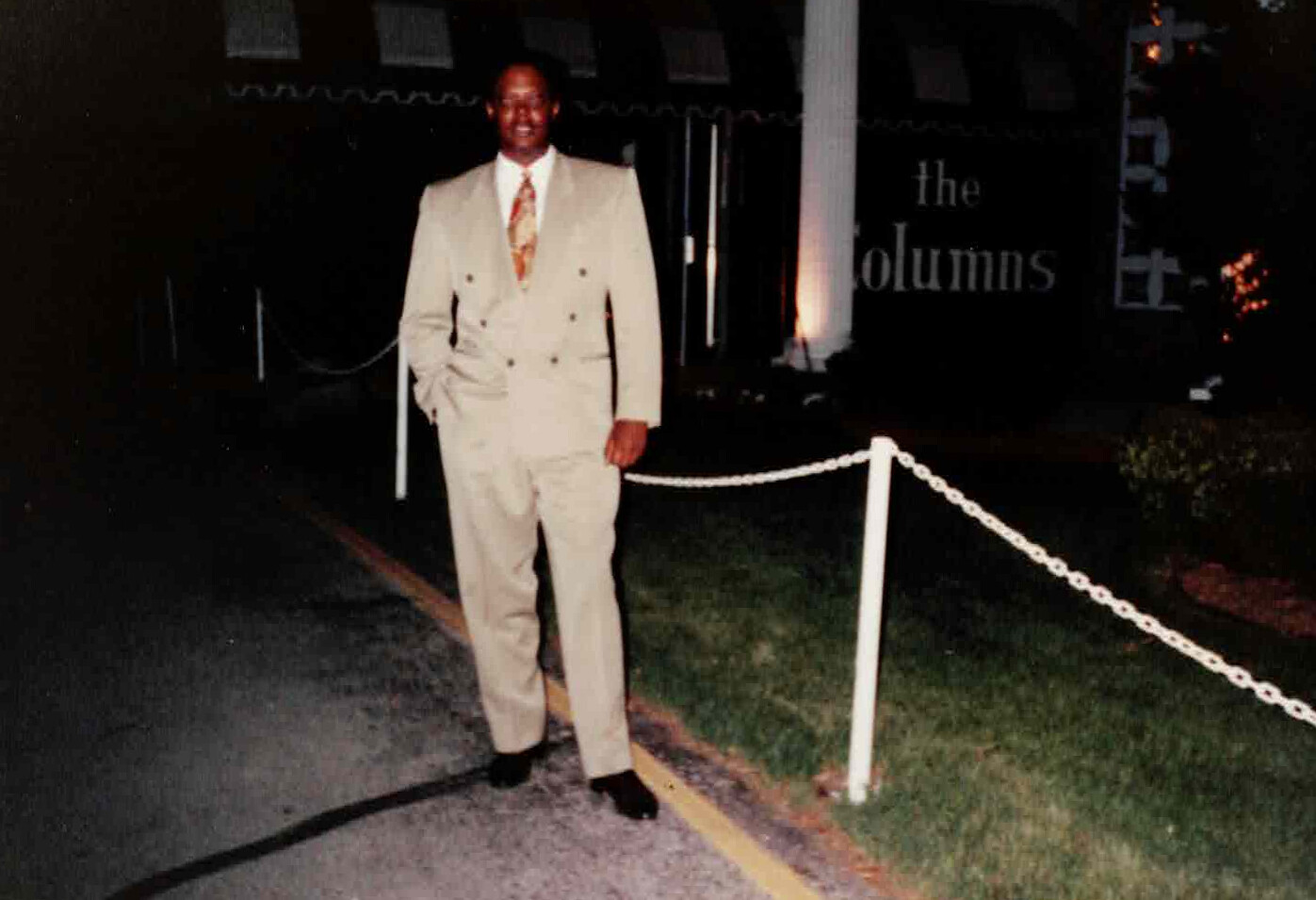 Man in a light beige double-breasted suit posing outdoors at night near a white rope barrier outside a building named The Columns.