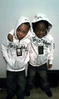 Two young children in matching white hooded jackets with black graphics, standing closely and looking at the camera.
