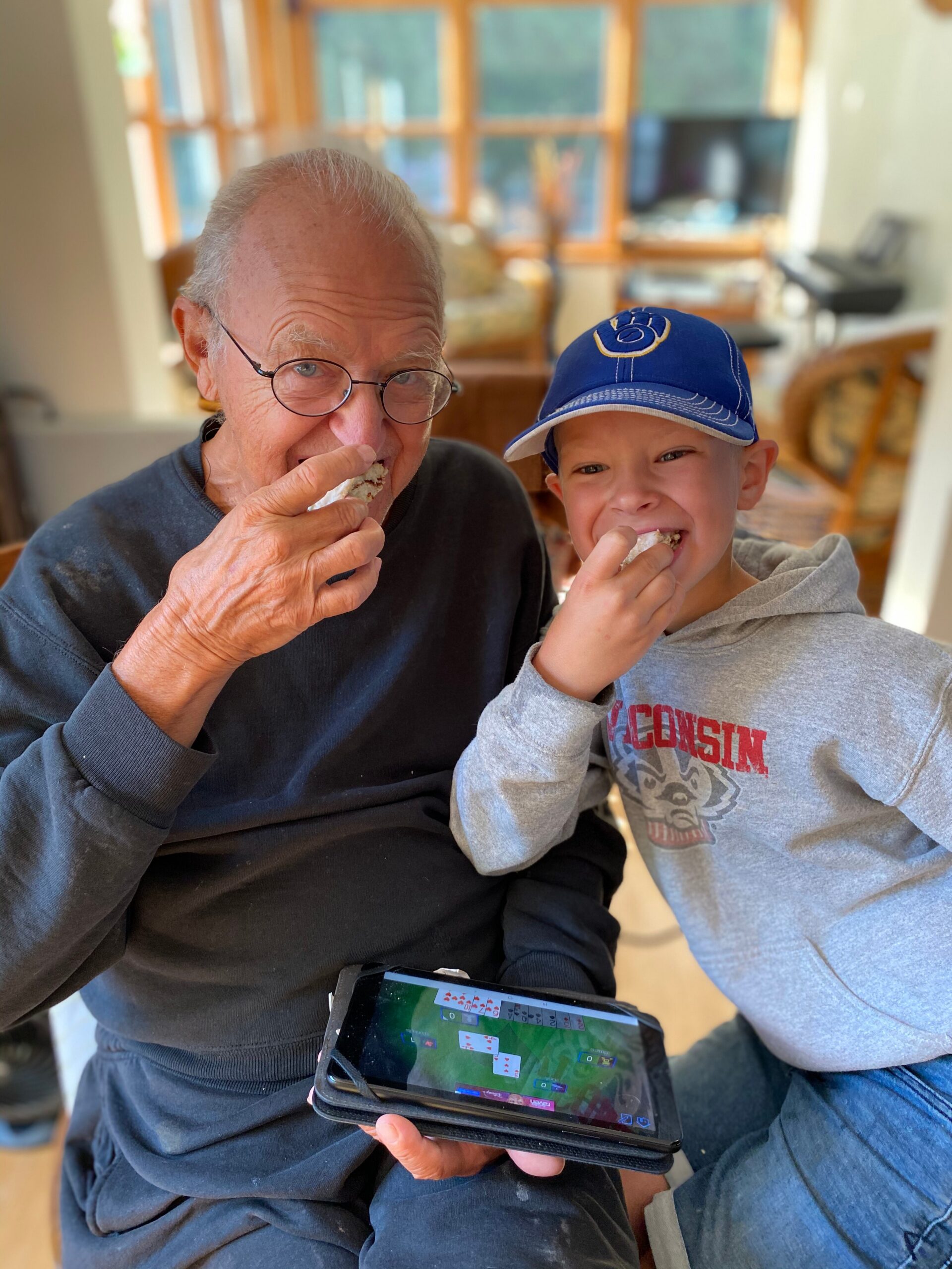 Older man with glasses and a smiling child eating a snack while looking at a tablet in a cozy living room setting.