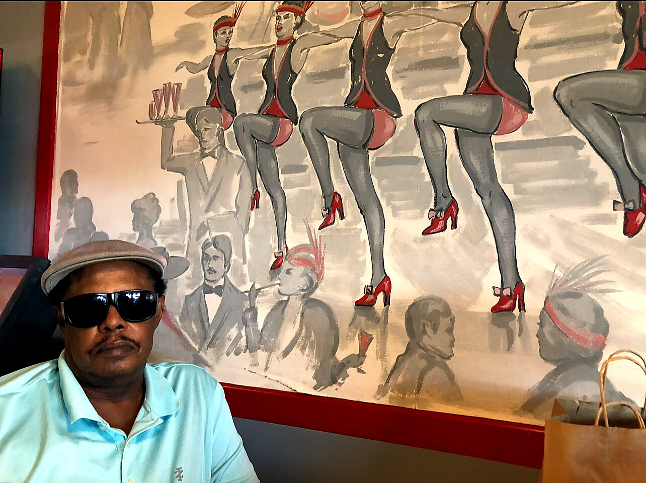 Man wearing sunglasses and a hat sits in front of a mural featuring line-dancers in red high heels and black outfits.