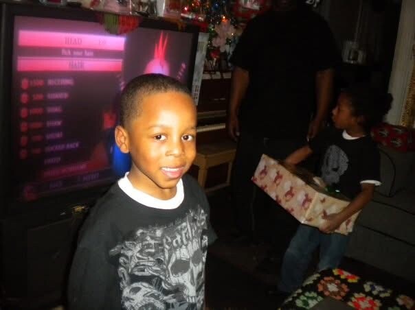 A smiling boy in a dark shirt stands near a TV in a holiday-lit living room with a man and another child in the background holding a gift box.