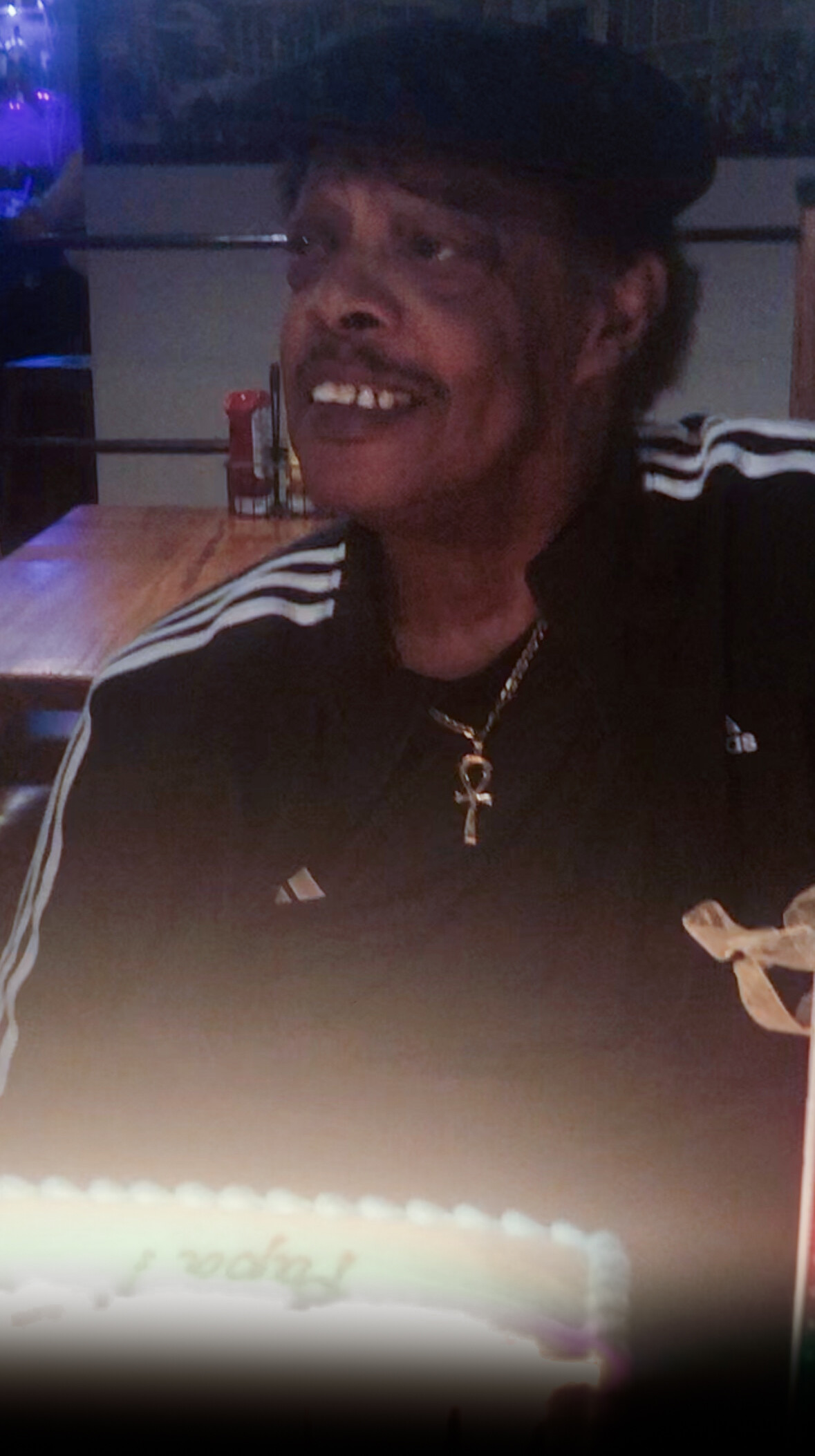 Smiling man wearing a black Adidas jacket with white stripes and a gold ankh necklace, seated at a dimly lit table in a party setting.